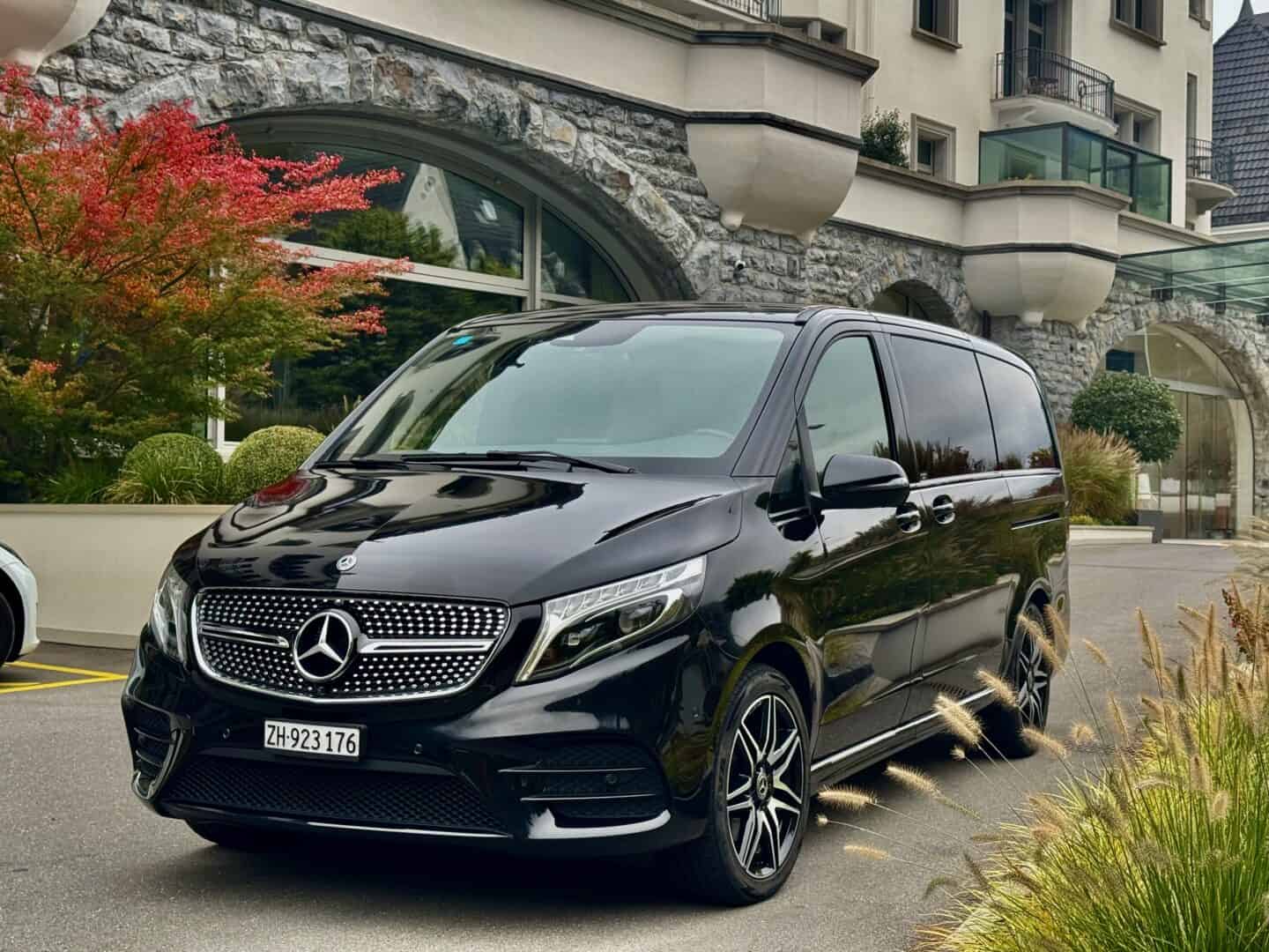 Black Mercedes-Benz V-Class luxury van parked in front of modern building with landscaped surroundings, ideal for corporate or private transportation services.