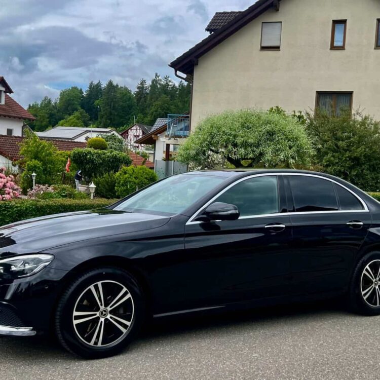 Luxury black sedan, Mercedes-Benz, parked on a quiet residential street in Switzerland, surrounded by greenery and houses, showcasing premium vehicle transportation services.