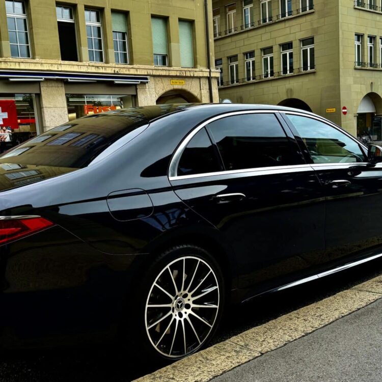 Luxury black sedan parked on city street with historic buildings and Swiss flag in the background.
