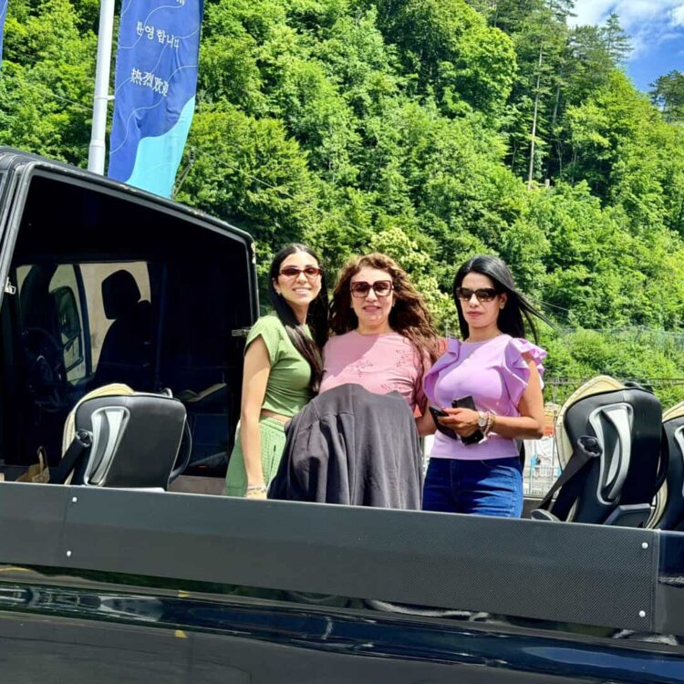 Three women standing in a black open-top vehicle with scenic green trees and blue sky in the background, representing reliable transport services.
